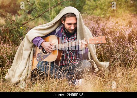 Joyeux jeune homme couvert de plaid jouant de la guitare acoustique et chantant la chanson. Homme en forêt de pins avec prairie de fleurs de bruyère. Gros plan, espace de copie. Banque D'Images