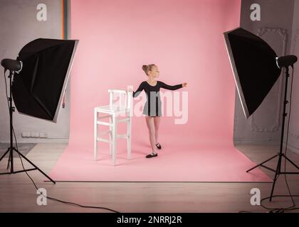 Portrait de jeune belle fille ballerina debout près de la chaise pratiquant le ballet vêtu noir robe de tutu posant dans le studio de photo fond rose Banque D'Images