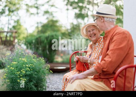 Femme âgée assise dans le jardin sur le banc avec son mari et tricoter le foulard rouge. Banque D'Images