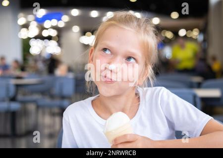 Portrait d'une petite fille drôle et affamée qui mange de la glace froide délicieuse dans une tasse de gaufres assis dans un café, fond sombre, bokeh. Un enfant aime la glace Banque D'Images