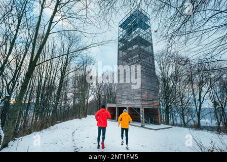 Deux hommes brillants et sportifs, vêtus de vêtements, qui traversent un parc d'arbres d'hiver, à côté d'une immense tour d'escaliers à l'architecture moderne. L'amitié et la santé des hommes Banque D'Images