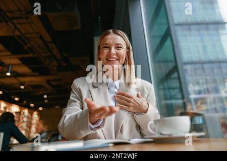 Femme d'affaires souriante communiquant en langage des signes tout en regardant l'appareil photo et assis dans un café Banque D'Images