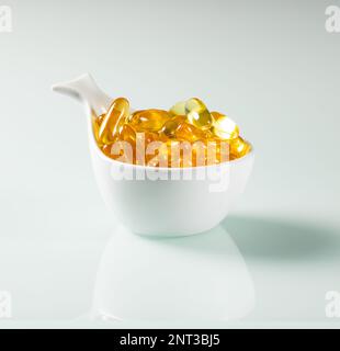 Une tasse blanche en forme de poisson remplie de capsules d'huile de poisson se reflète dans une surface en verre blanc. Banque D'Images