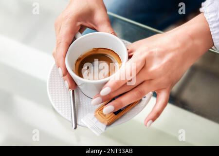 Tasse blanche avec café noir dans les mains d'une fille dans un café d'été. Une fille dans une chemise blanche et un Jean bleu tient une tasse de café blanc dans ses mains W Banque D'Images