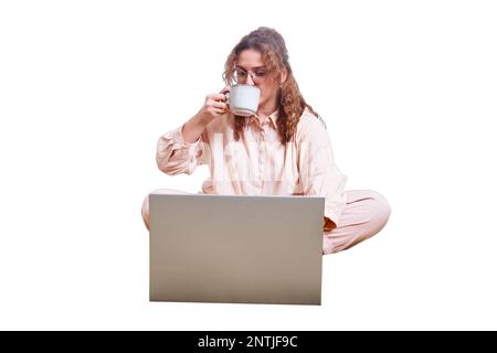 Une femme mignonne boit du café sur le lit de la maison, isolé sur un fond blanc. REDHEAD femme avec un ordinateur travaillant à distance pendant l'isolement en raison de quara Banque D'Images