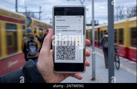 Berlin, Allemagne. 27th févr. 2023. Sur l'écran d'un smartphone, le code QR du Nationwide Deutschland-Ticket est affiché à côté des logos de VBB et BVG, tandis que deux trains S-Bahn sont stationnés à la gare S-Bahn en arrière-plan. Credit: Monika Skolimowska/dpa/Alay Live News Banque D'Images