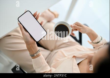 Magnifique et attrayant homme d'affaires asiatique sirotant un café tout en utilisant son smartphone à son bureau. maquette d'écran blanc de smartphone. vue de dessus Banque D'Images