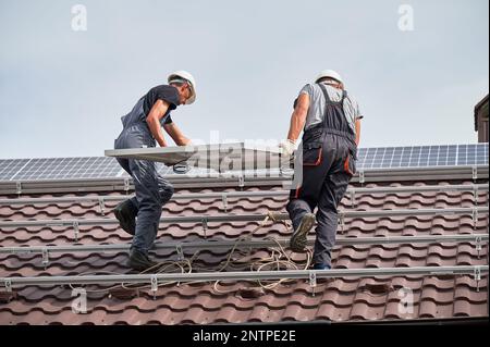 Hommes installateurs portant le module solaire photovoltaïque sur le toit de la maison. Électriciens dans les casques d'installation du système de panneaux solaires à l'extérieur. Concept d'énergie alternative et renouvelable. Banque D'Images