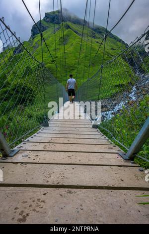 Un homme marche sur un pont suspendu entre les montagnes trop cultivées avec de l'herbe Banque D'Images