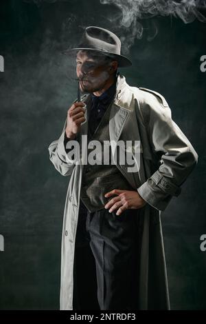 Portrait de l'homme, détective en trench-coat et chapeau fedora posant avec sérieux visage et fumant sur fond vert foncé vintage. Banque D'Images