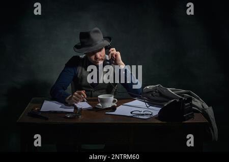 Détective professionnel en chapeau fedora assis à table et parlant au téléphone sur fond vert foncé vintage. Enquête Banque D'Images