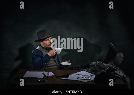 Détective professionnel dans le chapeau fedora assis à table avec le visage réfléchi et boire un café sur fond vert foncé vintage. Inspecteur Banque D'Images