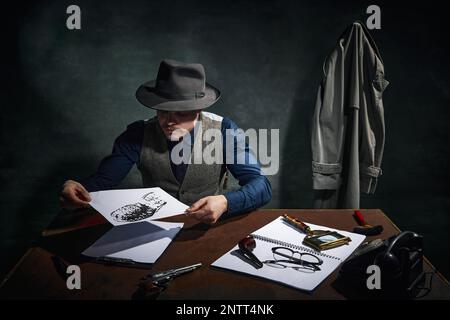 Détective professionnel en chapeau fedora assis à table et regardant l'empreinte, indice de crime sur fond vert foncé vintage. Enquête Banque D'Images