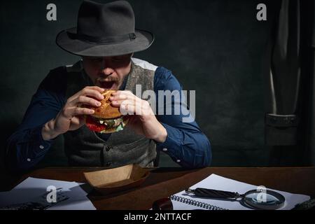 Détective professionnel dans le chapeau fedora assis à table et manger délicieux hamburger pour le déjeuner sur fond vert foncé vintage Banque D'Images