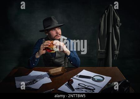 Détective professionnel dans le chapeau fedora assis à table et manger délicieux hamburger pour le déjeuner sur fond vert foncé vintage Banque D'Images
