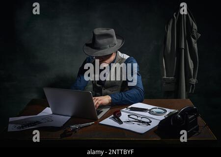 Détective professionnel en chapeau fedora assis à table et travaillant sur ordinateur portable sur fond vert foncé vintage. Enquête professionnelle Banque D'Images