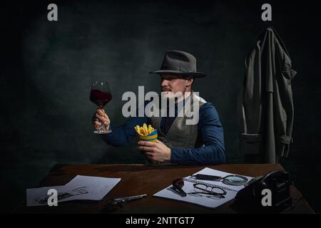 Détective professionnel dans le chapeau fedora assis à table, boire du vin rouge et manger des frites sur fond vert foncé vintage Banque D'Images