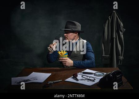 Détective professionnel dans le chapeau fedora assis à table, boire du vin rouge et manger des frites sur fond vert foncé vintage Banque D'Images