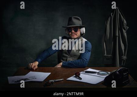 Homme élégant, détective professionnel en chapeau fedora et lunettes de soleil assis à la table et écouter des informations dans les écouteurs sur le vert foncé vintage Banque D'Images