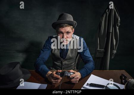 Détective professionnel en chapeau fedora assis à table et console de jeu sur fond vert foncé vintage Banque D'Images