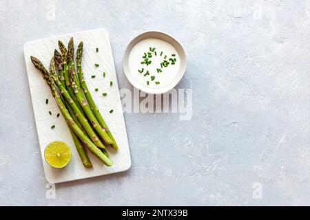 tiges d'asperges sur l'assiette rectangulaire servies avec du citron vert et du persil, fond en béton gris, vue de dessus, espace d'imitation Banque D'Images