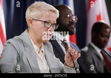 Femme blonde confiante en lunettes de vue et costume gris regardant le public pendant le discours tout en étant assise contre des collègues étrangers Banque D'Images
