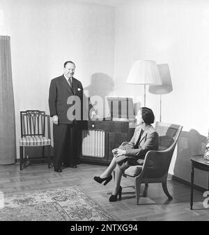 Gramophone dans l'ancien temps. Un homme âgé se tient fièrement devant un cabinet de gramophone où il a ouvert la couverture du gramophone et a placé un enregistrement de gramophone sur le plateau de gramophone. Sa femme est assise dans un fauteuil en osier dans leur maison bien rangée. À gauche du plateau gramophone se trouve une radio, d'où le nom radiogramophone, où les deux unités s'assoient ensemble dans un meuble. Suède 1954 Kristoffersson réf. BA100-12 Banque D'Images
