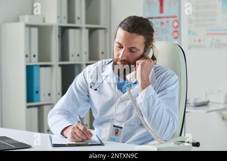 Jeune médecin avec récepteur téléphonique à l'oreille prenant des notes dans le document médical tout en écoutant le patient pendant l'appel téléphonique Banque D'Images