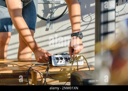 un homme méconnaissable, qui installe une session de musique en direct à l'extérieur, installe des câbles audio dans un contrôleur de mixage audio pour enregistrer et amplifier le son a. Banque D'Images