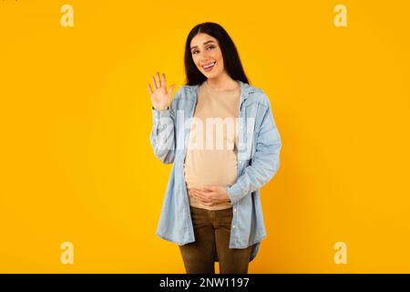 Belle femme enceinte touchant le ventre de la grossesse et en agitant en disant bonjour et souriant à l'appareil photo, fond jaune Banque D'Images