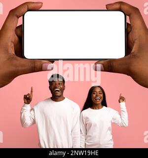 Joyeux couple noir pointant vers le smartphone Big Blank dans les mains des femmes géantes Banque D'Images