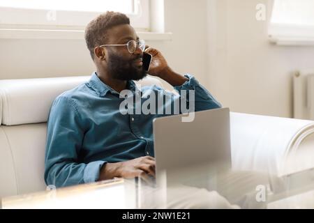 Homme d'affaires noir gai parlant sur smartphone avec clenit, assis sur un canapé avec ordinateur portable, travaillant dans un espace de bureau libre Banque D'Images