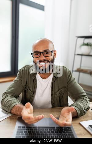 Positif latin barbu homme regardant l'écran d'ordinateur, ayant chat en ligne, gestuelle et souriant, assis au bureau Banque D'Images