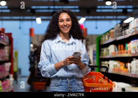 Shopping dans un supermarché. Une jeune femme latino-américaine se tient entre des rangées d'étagères avec un chariot. Il tient le téléphone, a une liste, écrit des messages, commande et paie en ligne. Sourire à l'appareil photo. Banque D'Images