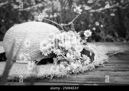 Bouquet de fleurs sauvages avec chapeau de paille sur une surface en bois à l'extérieur. Ton noir et blanc Banque D'Images