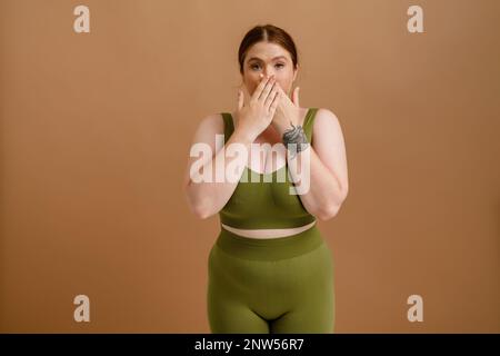 Jeune femme blanche aux cheveux de gingembre posant tout en couvrant sa bouche isolée sur fond beige Banque D'Images