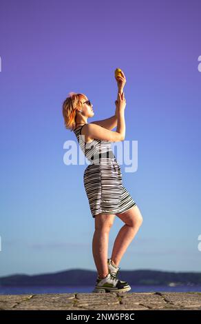 Jeune femme debout sur le bord de la mer pose statue avec citron et robe à rayures Banque D'Images