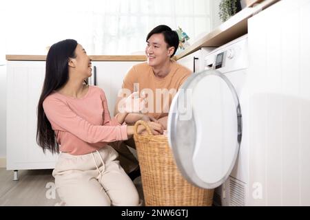 Les jeunes Asiatiques se sont mariés heureux couple souriant et faisant la lessive à la maison. Petit ami et petite amie mettant les vêtements dans la machine à laver à chargement frontal ensemble Banque D'Images