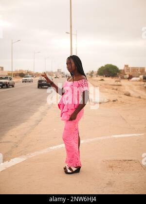 Une jeune mauritanienne vêtue d'une robe rose de mode africaine hante un taxi dans la rue. Banque D'Images
