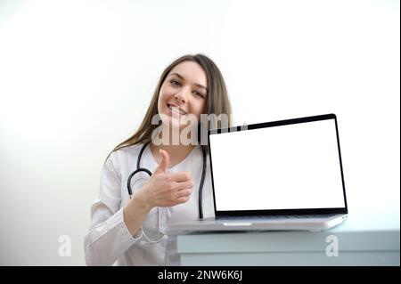 jeune fille médecin infirmière s'assoit près d'un ordinateur portable et montre pouce vers le haut la publicité d'un produit sur un fond blanc texte de la publicité des suppléments alimentaires bonne nutrition mode de vie sain sourire tendresse Banque D'Images