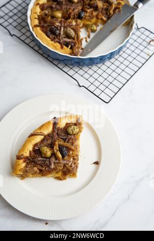 Tarte aux oignons Pissaladiere avec anchois et olives noires. Le morceau de tarte découpé repose sur la plaque, le reste du moule en arrière-plan Banque D'Images