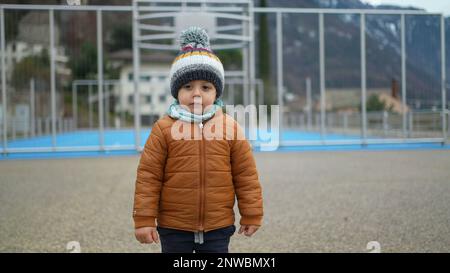 Un petit garçon se tient au parc pendant la saison hivernale, portant un foulard et une veste. Un enfant en bas âge pensif se tient à l'extérieur avec des vêtements chauds Banque D'Images