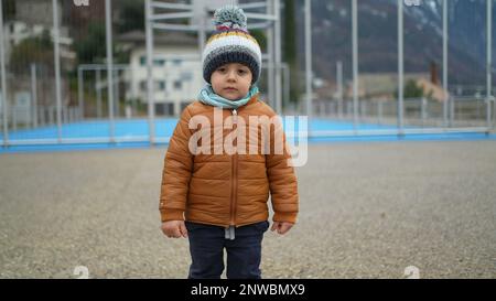 Un petit garçon se tient au parc pendant la saison hivernale, portant un foulard et une veste. Un enfant en bas âge pensif se tient à l'extérieur avec des vêtements chauds Banque D'Images