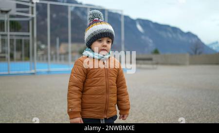 Un petit garçon se tient au parc pendant la saison hivernale, portant un foulard et une veste. Un enfant en bas âge pensif se tient à l'extérieur avec des vêtements chauds Banque D'Images