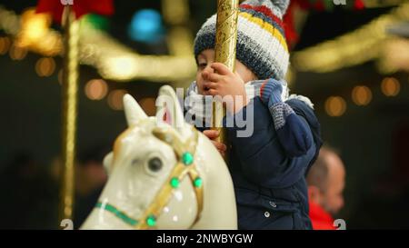 Un joyeux petit garçon qui fait du carrousel en mouvement et qui s'enorme bonjour. Un enfant en merry se déplace au parc d'attractions en portant un bonnet et un foulard pendant l'hiver Banque D'Images