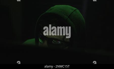 Hacker porte des lunettes noires en train de taper sur l'ordinateur dans une pièce à faible éclairage avec un éclairage vert étrange Banque D'Images