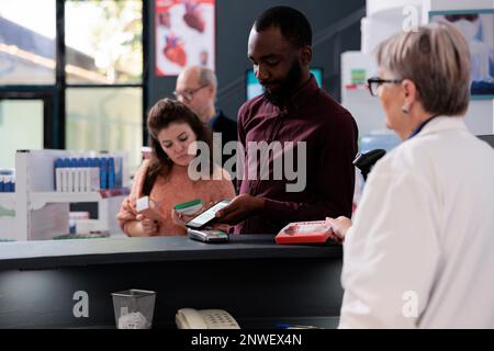 Jeune client adulte mettant smartphone sur pos terminal payer pour des suppléments en pharmacie après avoir discuté de traitement de santé avec le pharmacien. Client de pharmacie acheter des pilules de prescription Banque D'Images
