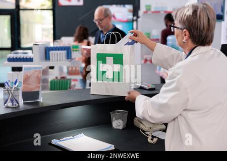 Pharmacien âgé debout au comptoir de la pharmacie mettant le traitement d'ordonnance dans un sac en papier préparant le colis pour le client. Les clients regardant les étagères pleines de médicaments, achetant des pilules Banque D'Images
