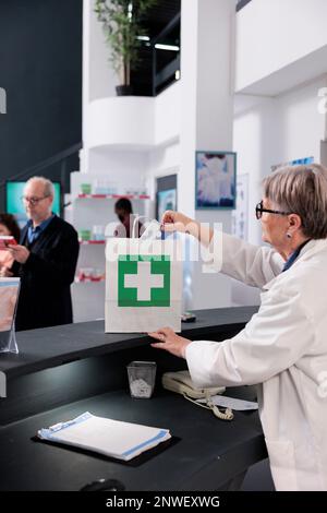 Pharmacien âgé debout au comptoir de pharmacie mettant des paquets de pilules dans un sac en papier en attendant que le client vienne payer pour le traitement sur ordonnance. Services et soutien en matière de soins de santé Banque D'Images
