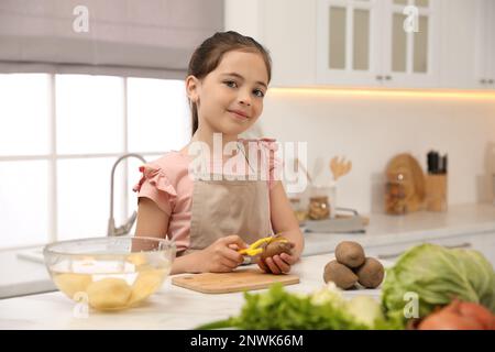 Petite fille épluchant la pomme de terre à la table dans la cuisine. Préparation des légumes Banque D'Images